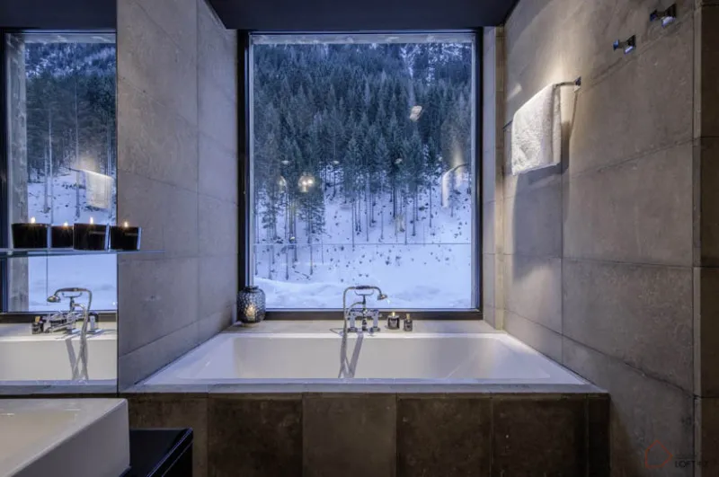 A modern concrete bathroom with a large window overlooking a snowy forest.