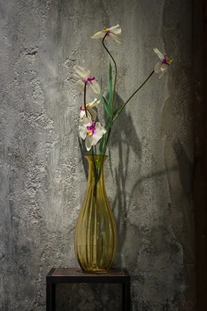An industrial-style vase filled with flowers on a black table against a textured wall background.