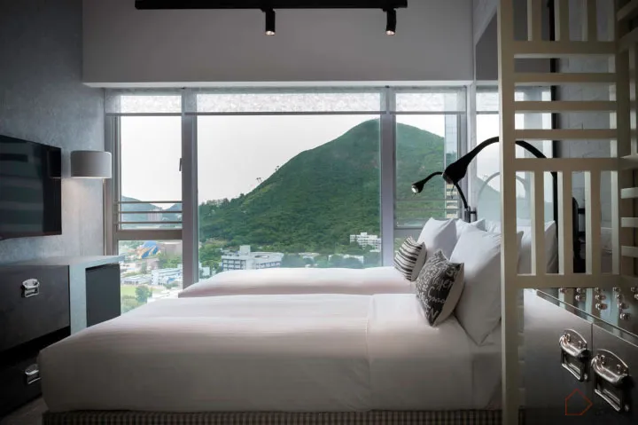 Modern hotel room featuring a bed with white bedding and a large glass window overlooking a green mountain.