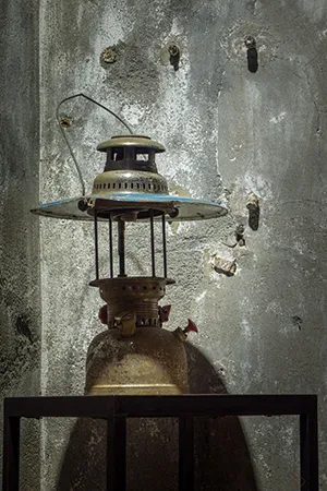 An antique, metal lantern with a glass chimney and handle, placed on a wooden shelf against a worn, concrete wall.
