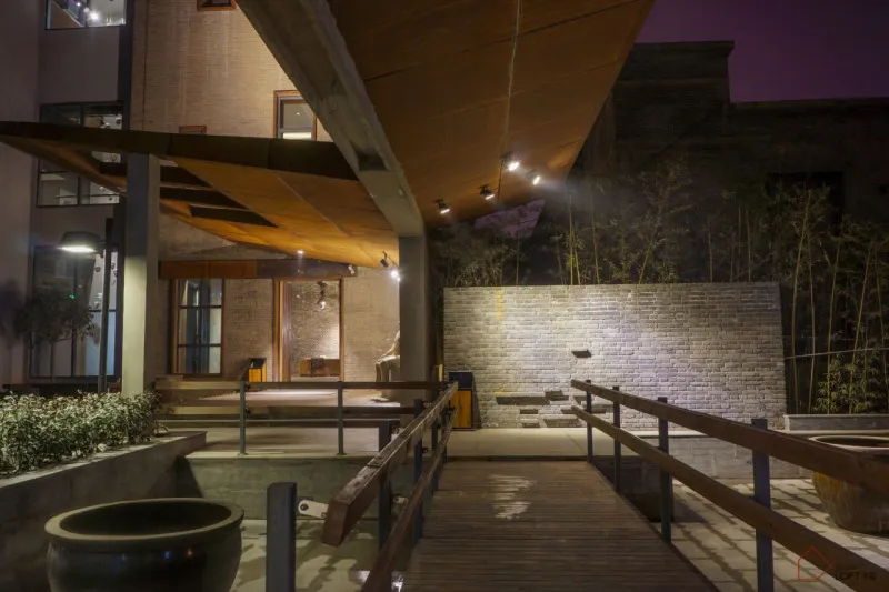 Urban scene with wooden walkway, brick wall, and concrete patio illuminated with urban lighting.