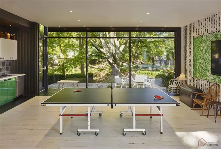 A modern game room featuring a wooden ping pong table, white chairs, and a green wall with a leaf pattern.