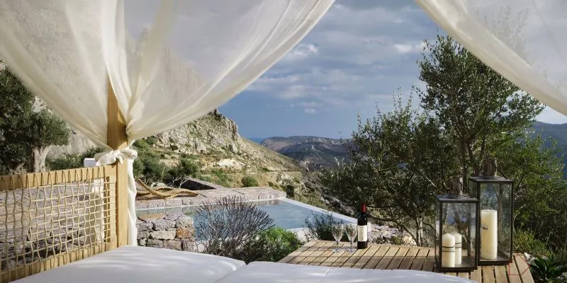 Luxurious poolside lounging area with textured fabric canopy, wooden deck, stone walls, glass drinkware, and scenic mountain