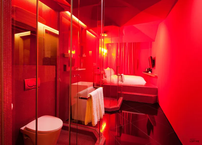 A modern bedroom with red lighting, glass partitions, concrete floor, mirrored walls, and fabric bedding.