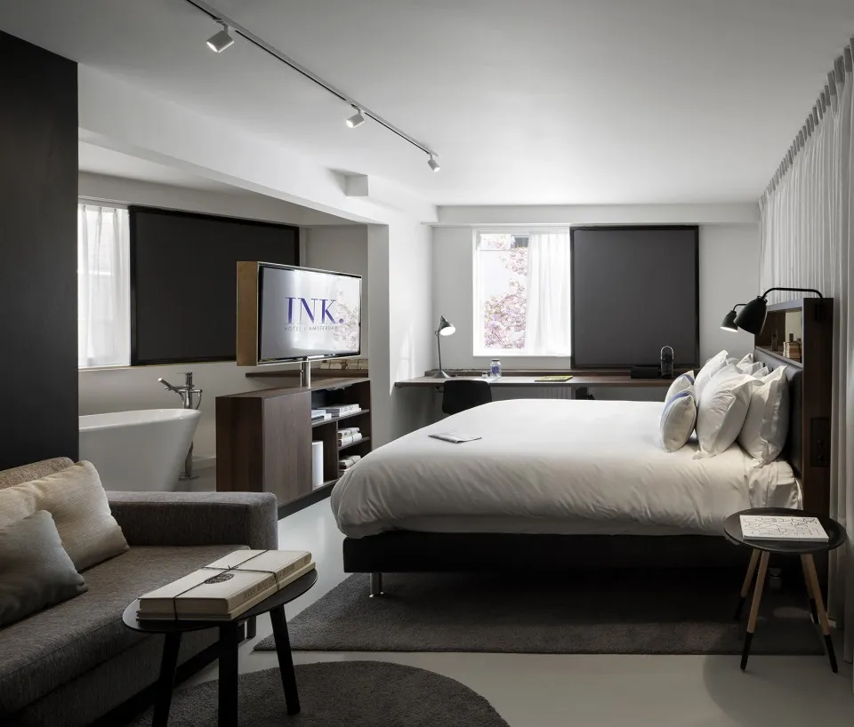 A modern minimalist hotel room with a bed, sofa, bathtub, and television.