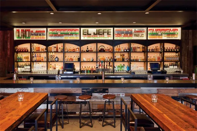 A spacious, modern restaurant bar featuring industrial-style lighting and a wide selection of drinks.