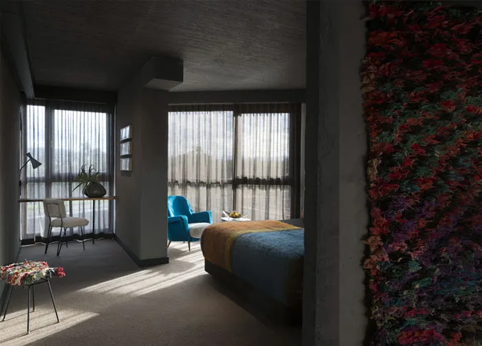 A modern bedroom featuring a concrete wall with embedded hyphae, a blue armchair, and a desk with a plant.