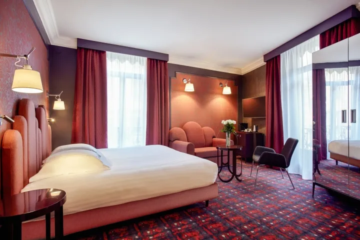 A luxurious hotel room featuring a bed, armchair, and desk with a television, all adorned with red and brown furnishings and