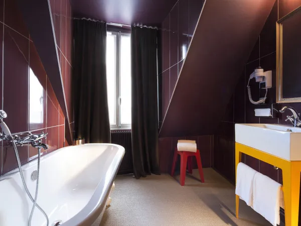 A modern bathroom with a white bathtub, brown tiles, and red stool.
