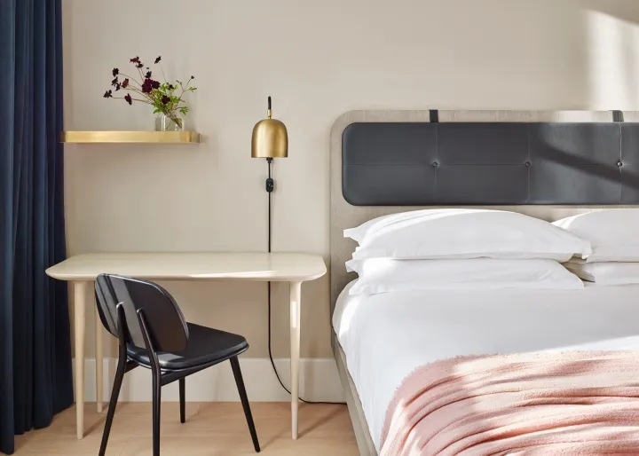 A modern minimalist bedroom featuring white bedding, a wooden desk, a black chair, a brass wall lamp, and a shelf with flower