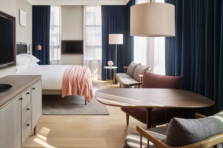 A cozy modern bedroom with a bed dressed in soft pink bedding, a wooden table, and a plush sofa adorned with cushions.