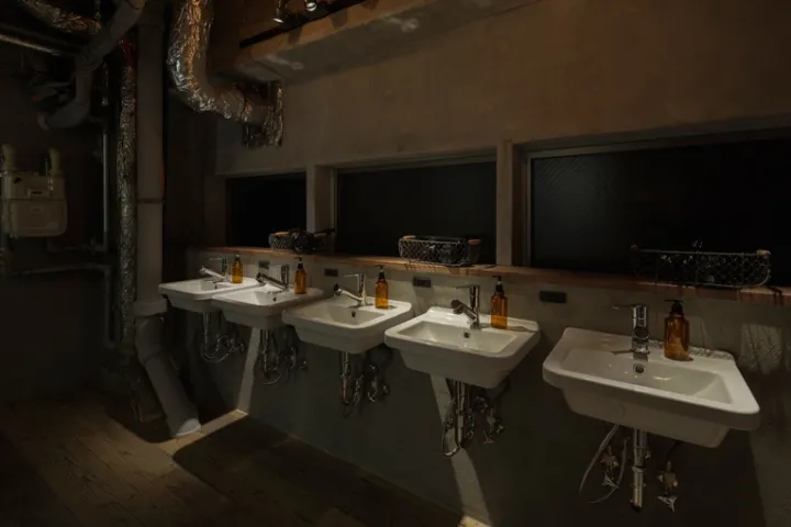 Multiple white basins with orange soap dispensers in a dimly lit, industrial-style public bathroom with concrete walls and ex