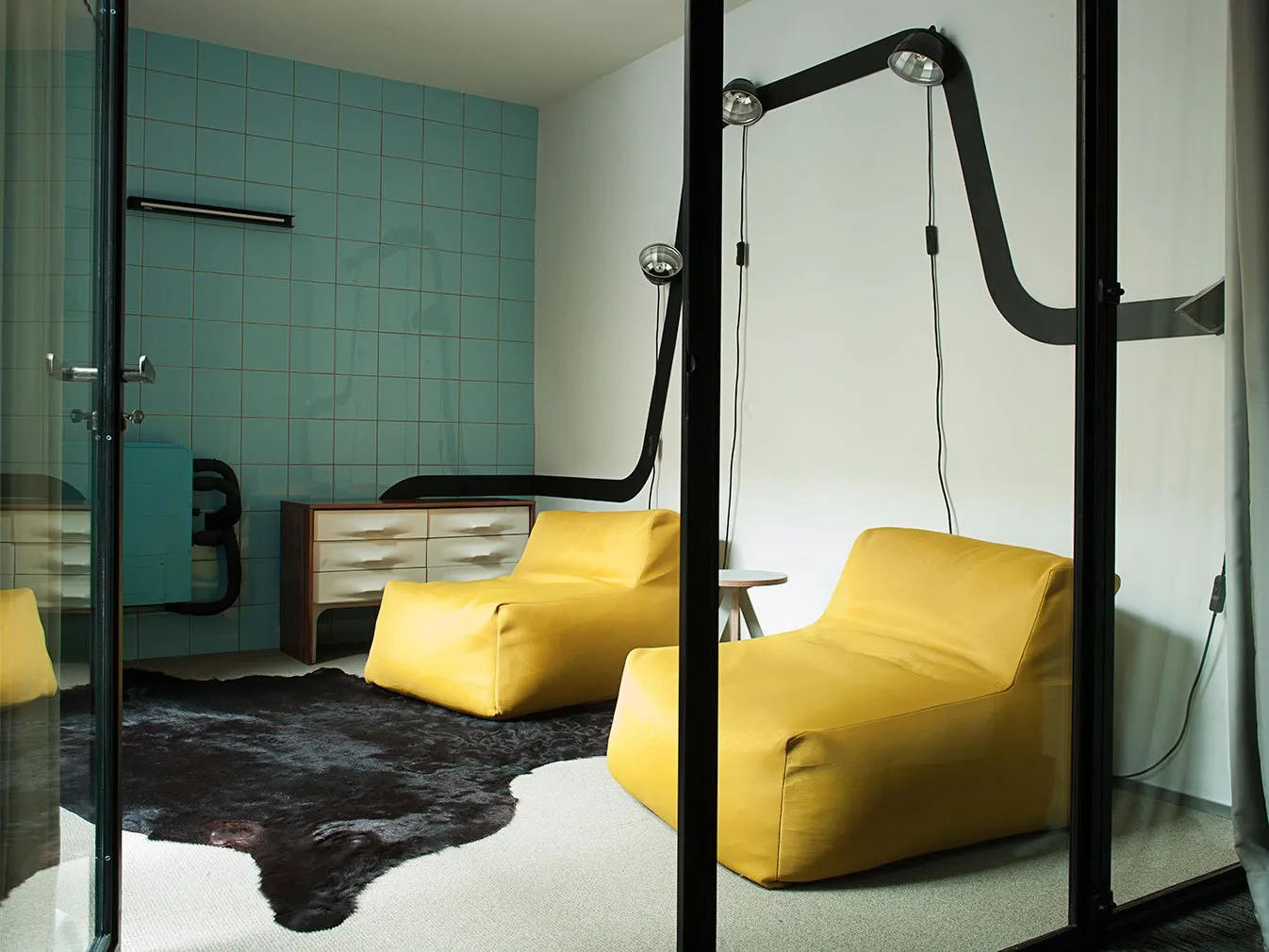 A minimalist living room with yellow fabric and leather chairs, white walls, and glass walls, featuring a cowhide rug.