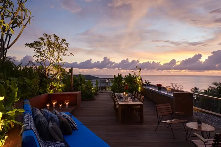 A rooftop terrace with wooden flooring, greenery, and sunset view.