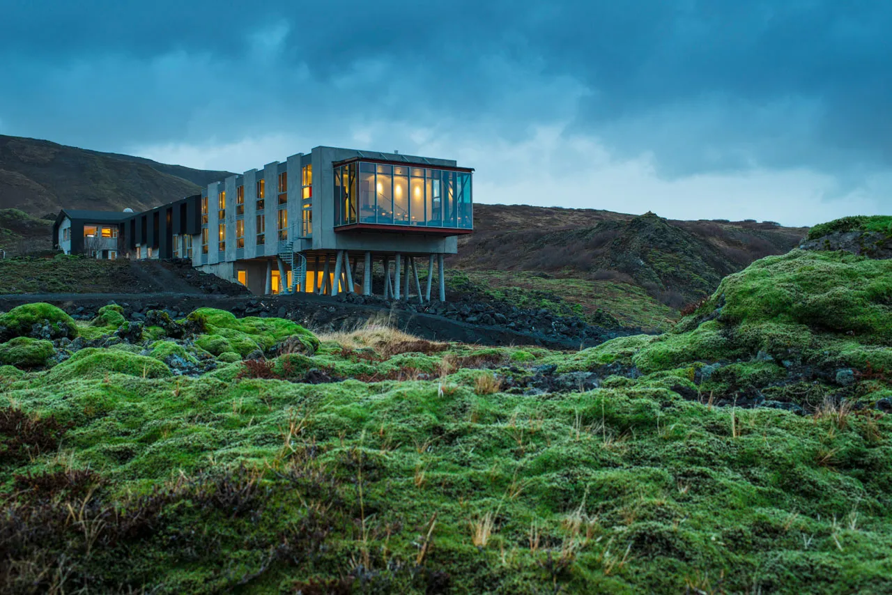A modern residential house with concrete exterior and large glass windows, surrounded by lush green moss-covered land.