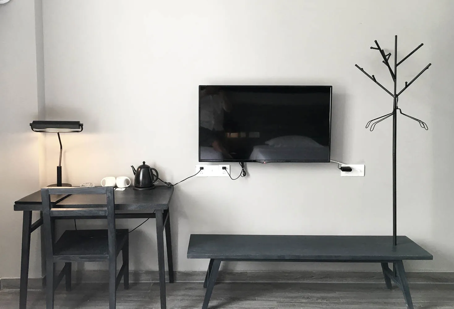 A minimalistic hotel room with a black desk, black bench, and a mounted television on the wall.