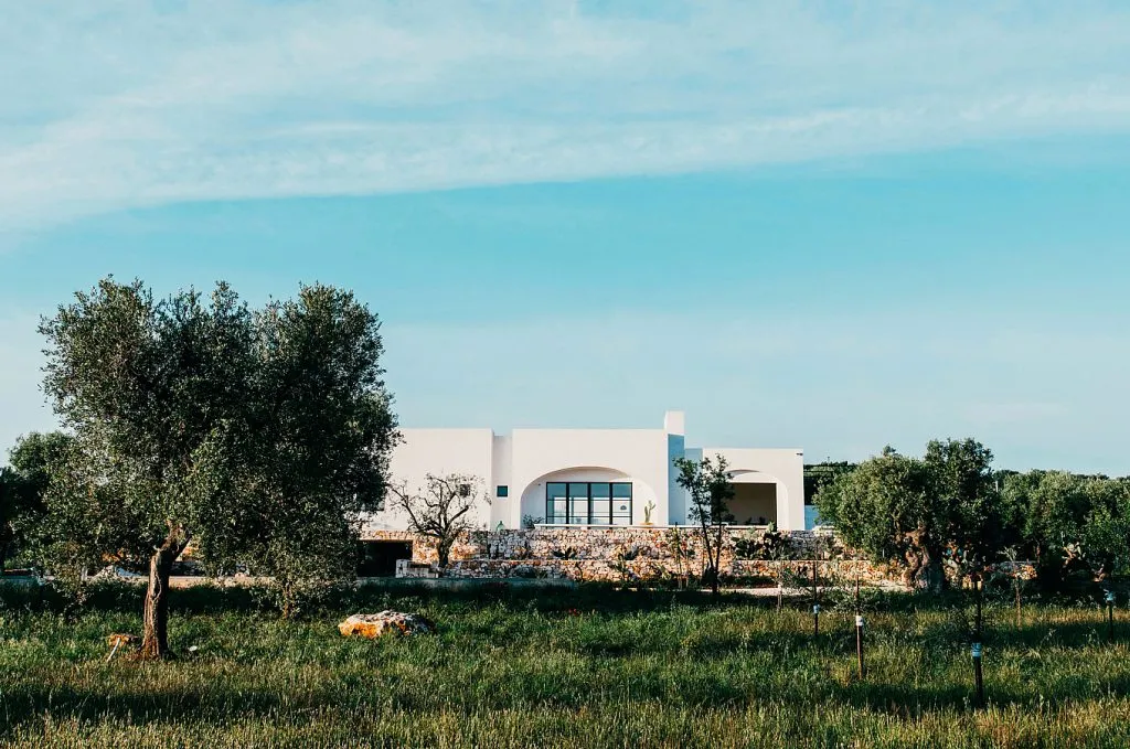 A modern residential farmhouse painted in white with a stone wall and green grass in the foreground.