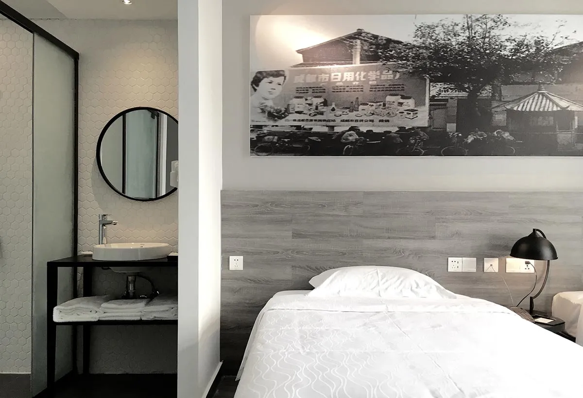 A minimalist hotel room with a single bed, a sink, and a framed black and white photograph of a street scene hanging on the w