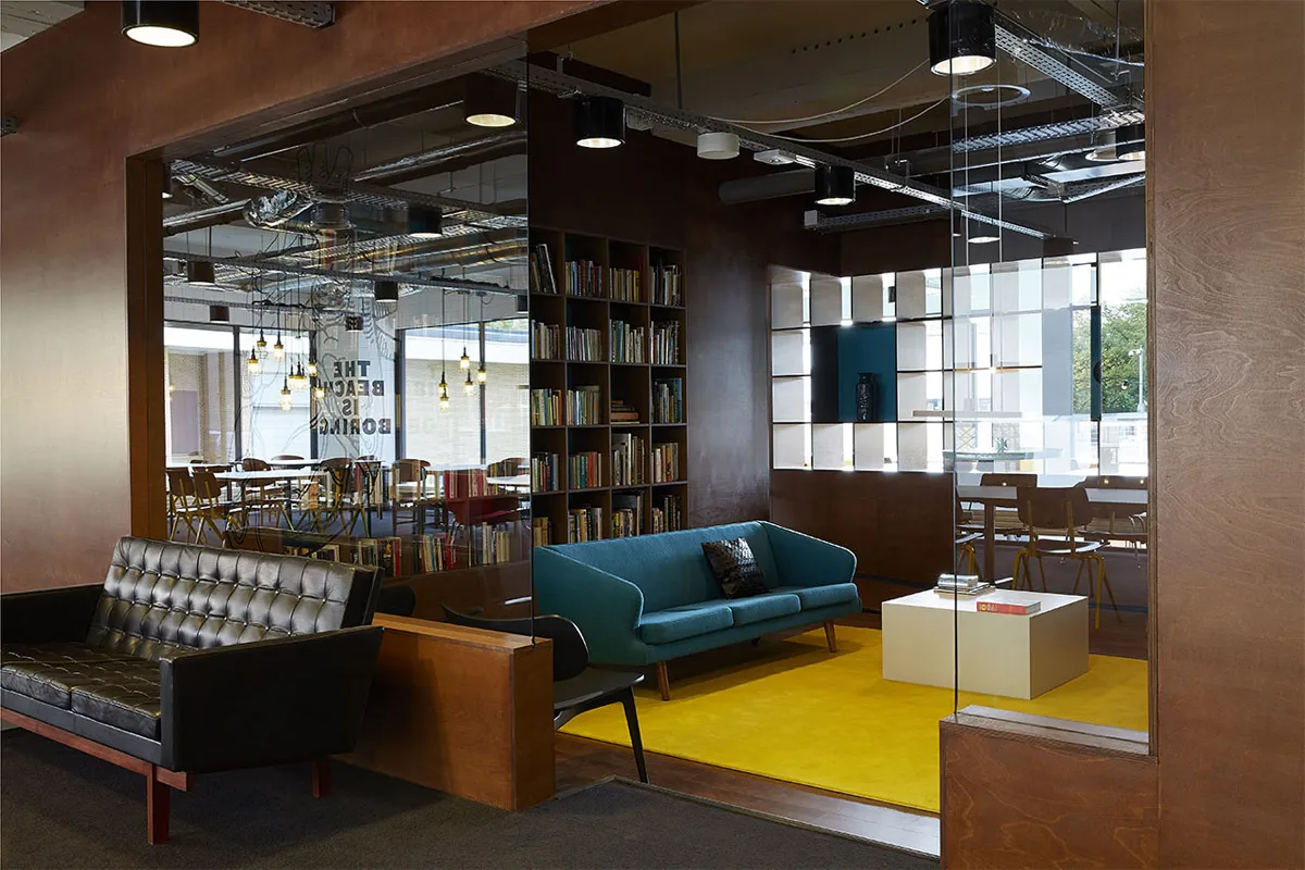 A modern office space featuring metal-framed furniture, with a mix of seating options, a bookshelf, and large windows.