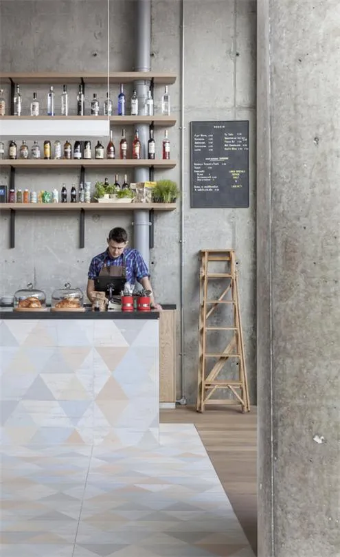 A worker stands behind a coffee bar adorned with various bottles and plants, with a menu chalkboard and a wooden step ladder 