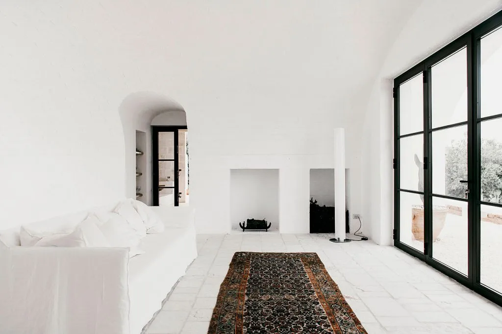 A minimalist living room featuring a concrete floor and walls, with a white sofa, a patterned rug, and a fireplace.