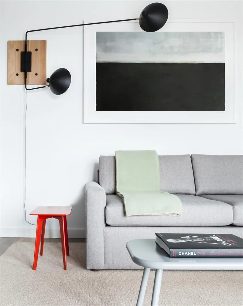 A modern grey sofa with a green throw blanket, white walls, and black floor lamp, accompanied by a red stool.