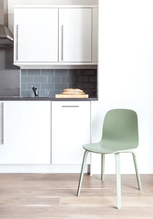 A modern minimalist kitchen with white cabinets, a green chair, and a wooden countertop. The kitchen features a white backspl