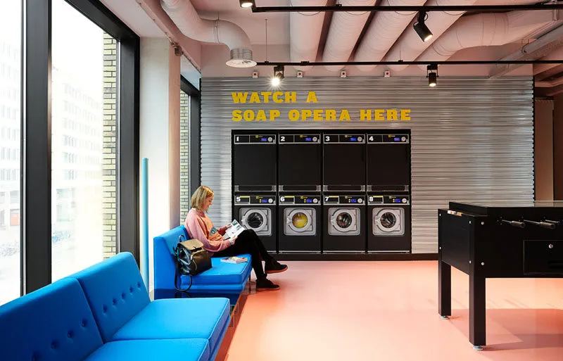 A person sitting on a blue cushion in a modern industrial laundromat lounge with concrete flooring and washing machines in th