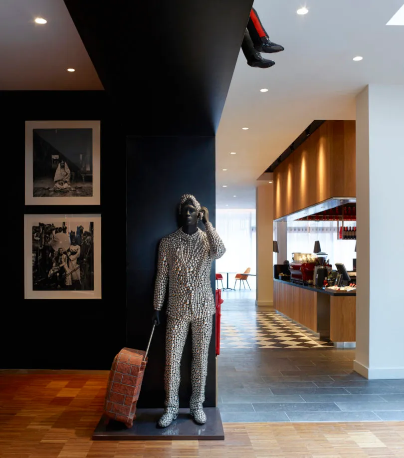 A modern statue of a person dressed in a patterned suit and holding a suitcase stands in a lobby with black and white checker