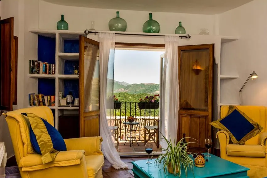 A cozy living room with vibrant yellow furniture and green accents, featuring a balcony with a view of lush mountains.