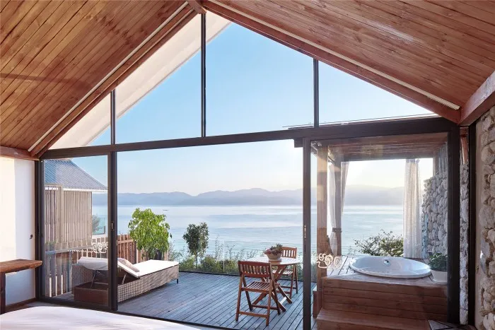 A spacious residential interior featuring a wooden deck overlooking the sea, stone wall, and glass wall and window.