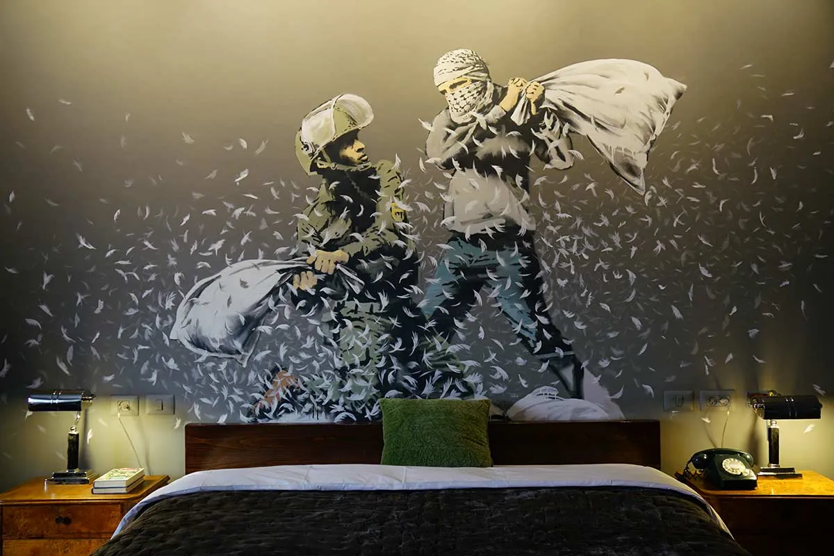 A black and white mural of two individuals engaged in a pillow fight on a bed in a hotel room. The wall features a bird feath