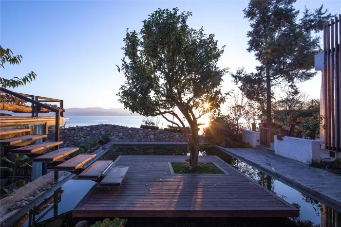 Outdoor scene featuring a wooden deck with a small tree in the center, surrounded by trees, a pool, and water in the backgrou