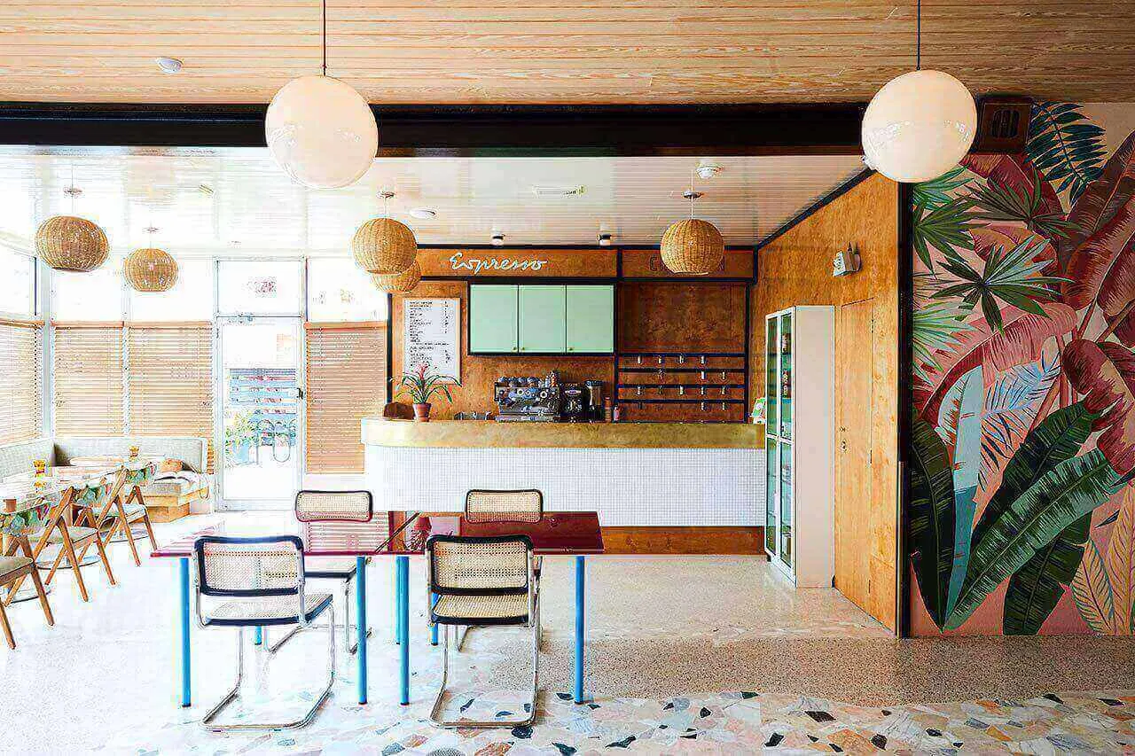 A modern cafe interior with a wooden bar and terrazzo floor, featuring wicker pendant lights and tropical mural.