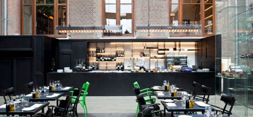 Interior design of a modern restaurant featuring a bar area with black countertops, wine bottles, and green chairs.