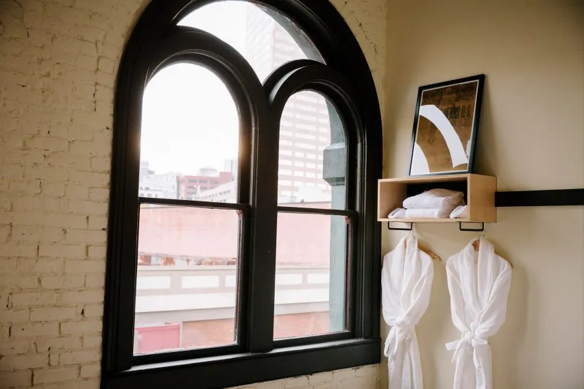 A hotel room with a white brick wall, black-framed window overlooking a cityscape, and two white robes hanging from hooks.