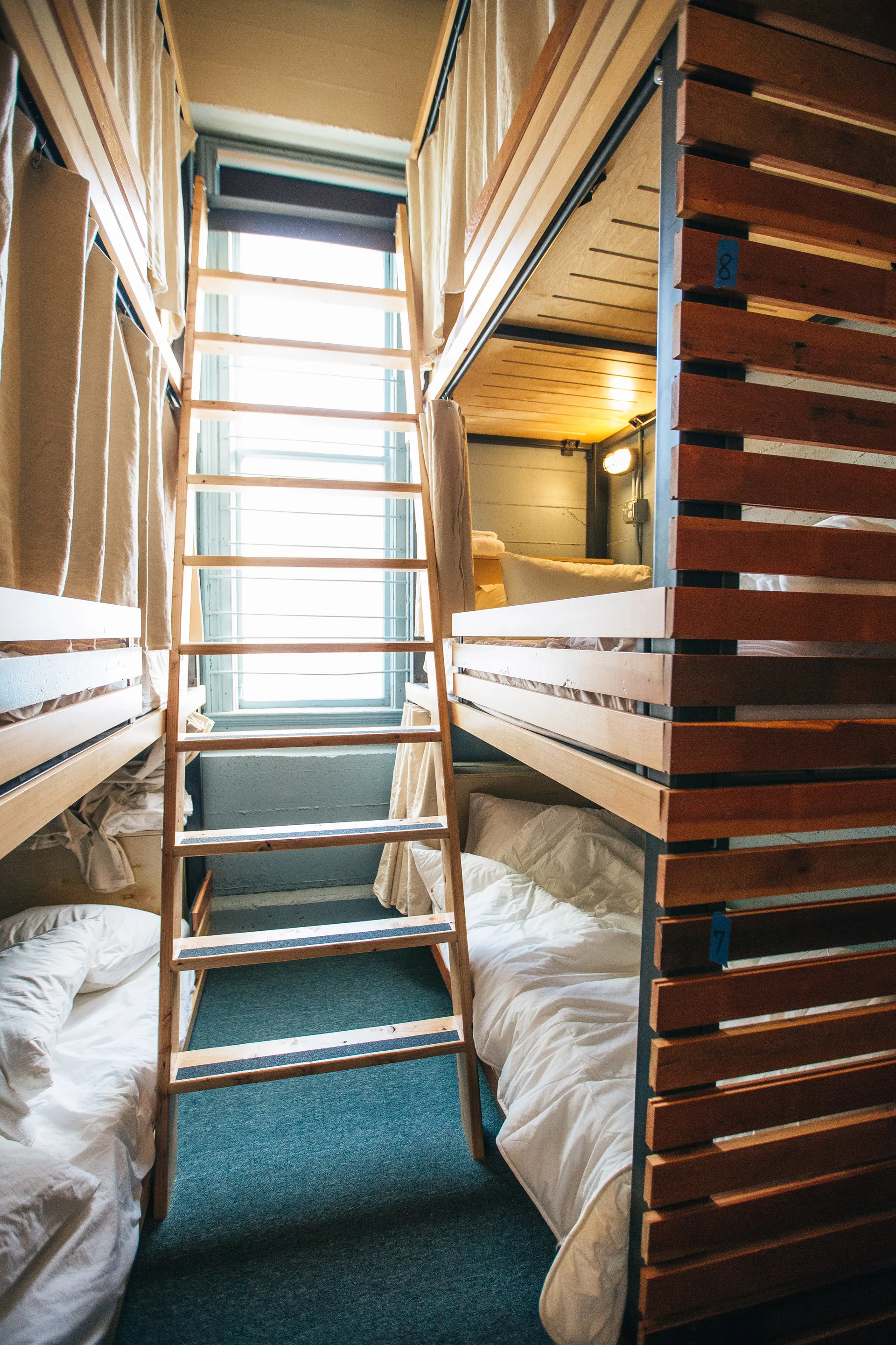 A hostel room with wooden bunk beds, metal ladder, and fabric curtains.
