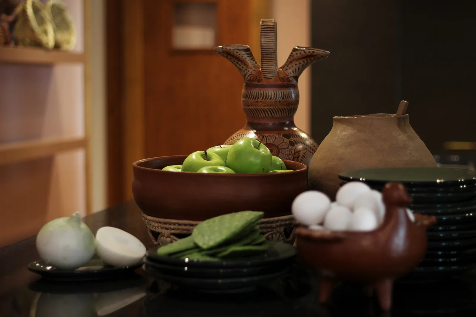 A still life arrangement featuring green apples in a bowl, eggs, sliced onions, and various ceramic bowls and dishes.