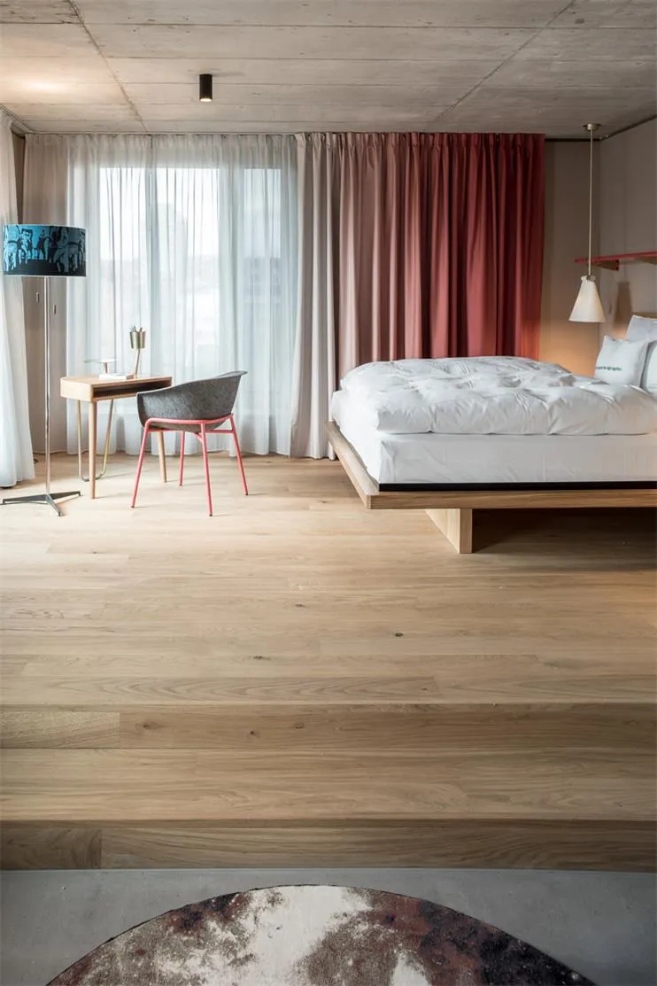 A modern bedroom featuring a wooden floor, a comfortable bed with a white mattress, and a desk area with a chair and a lamp.
