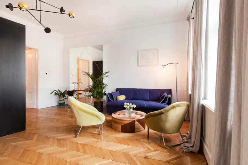 A cozy living room with wooden flooring, a blue sofa, yellow armchairs, and potted plants.