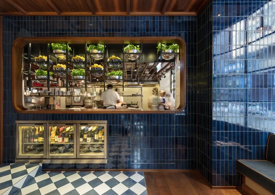 A restaurant kitchen with blue tiled walls, checkered floor, and open shelving displaying herbs and fruits.