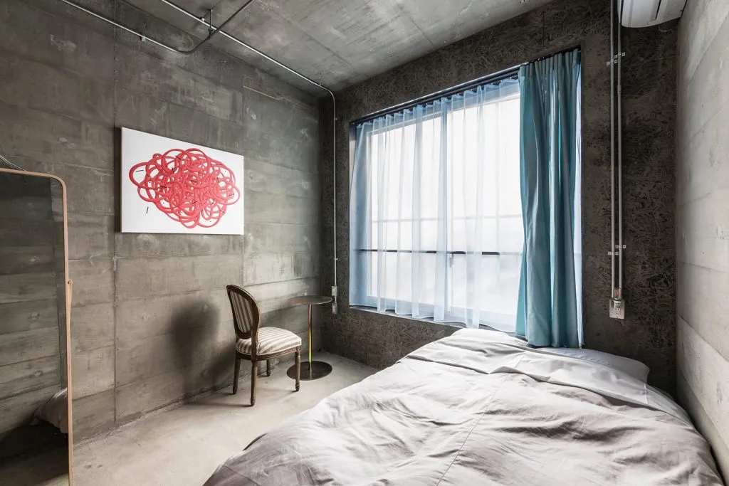 A minimalist bedroom featuring concrete walls, a bed with grey bedding, and a small table with a red and white abstract paint
