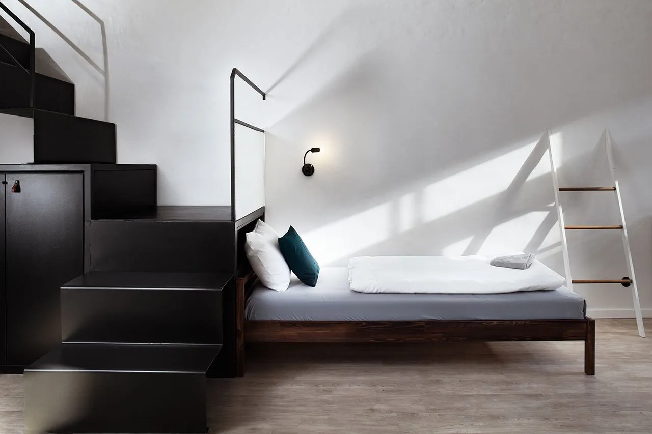 Minimalist bedroom with a wooden full-length bed, white bedding, and a black staircase leading to a cabinet.