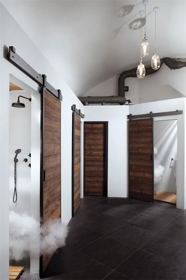 Industrial bathroom with wooden sliding doors and ceramic shower stalls, white walls and flooring, and exposed black pipes