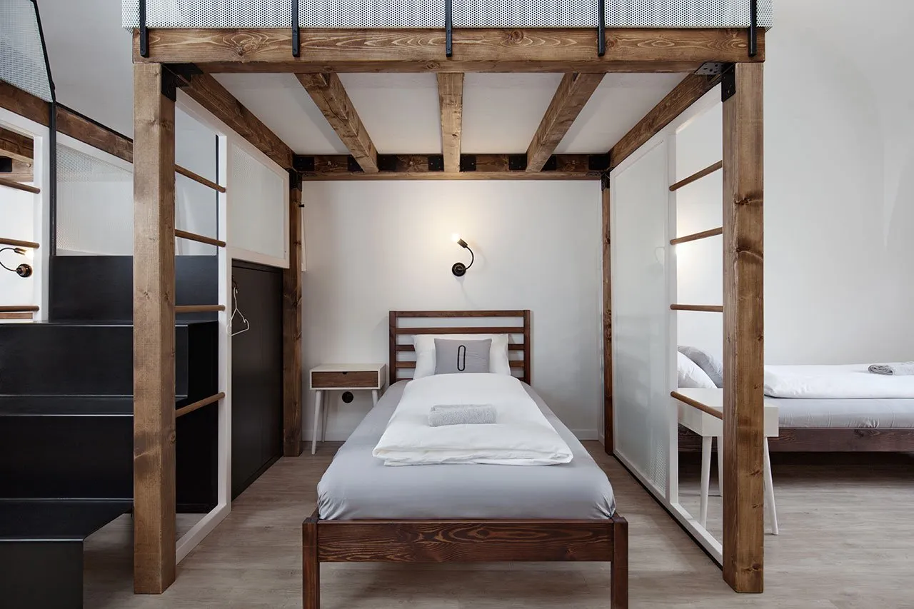 A minimalist bedroom with a single bed on a raised wooden frame, surrounded by transparent glass walls and metal railings.