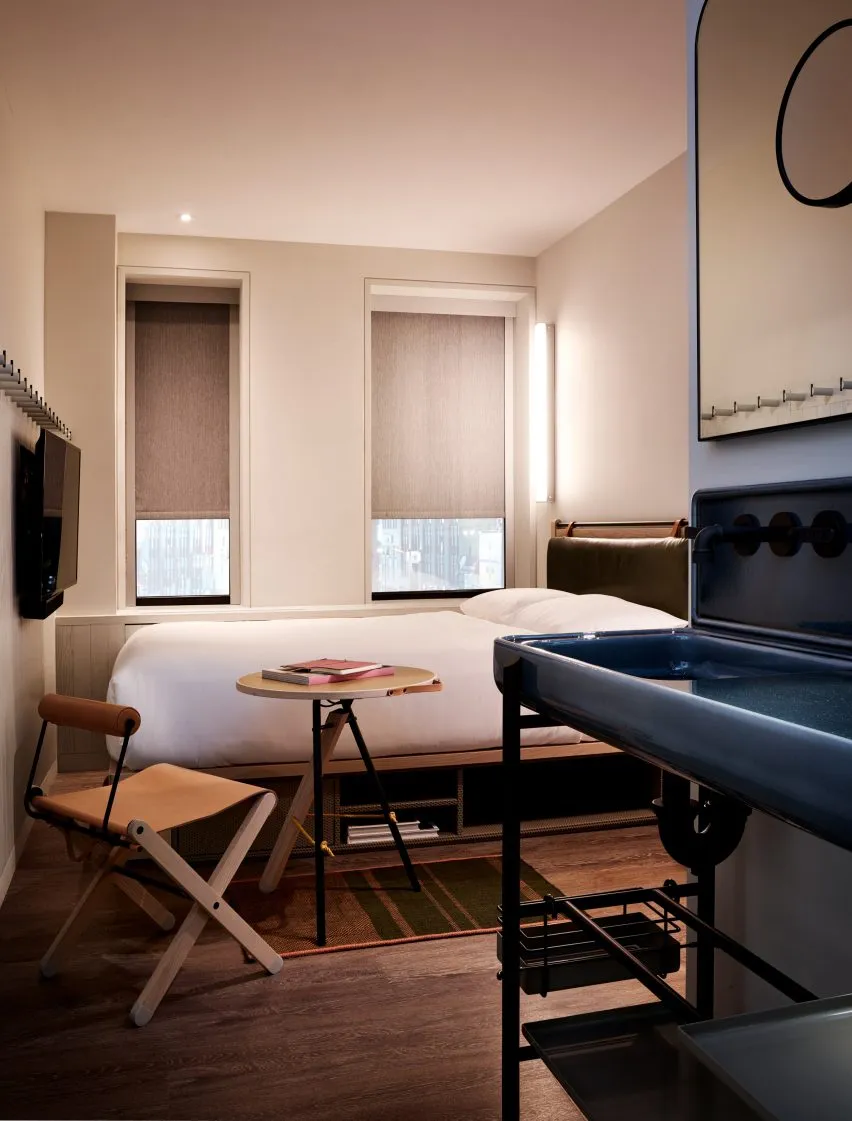 Minimalist bedroom featuring a bed with wooden and fabric elements, a small table with a book and glasses, and a metal sink.