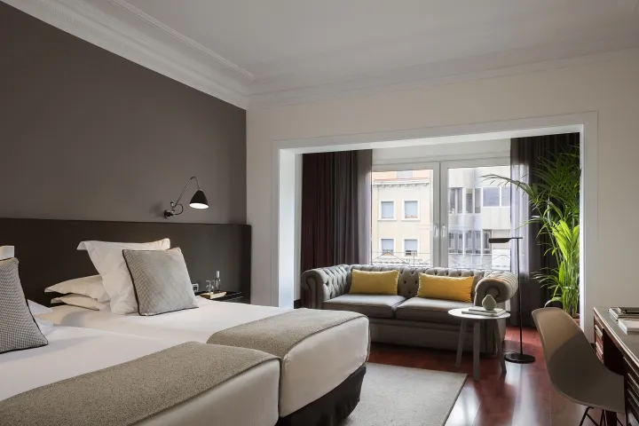 Modern, minimalist hotel suite with grey decor, featuring a comfortable seating area, a large bed with plush bedding, and a s