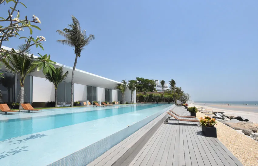 Modern glass and concrete beach resort with poolside palm trees and rock formations.