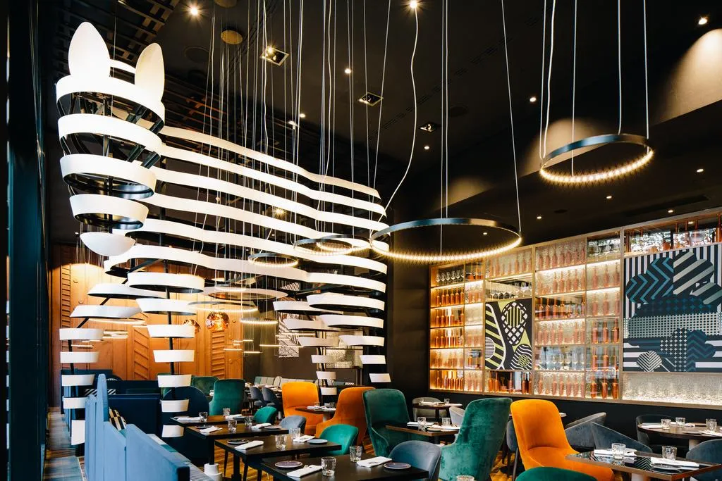 A modern restaurant interior with hanging lights, colorful chairs, and glass-fronted shelves.