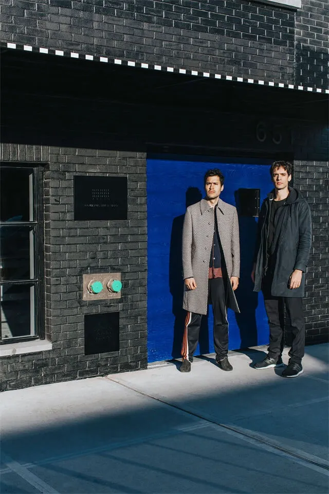 Two men standing in front of a black brick building with a blue garage door, wearing stylish clothing.